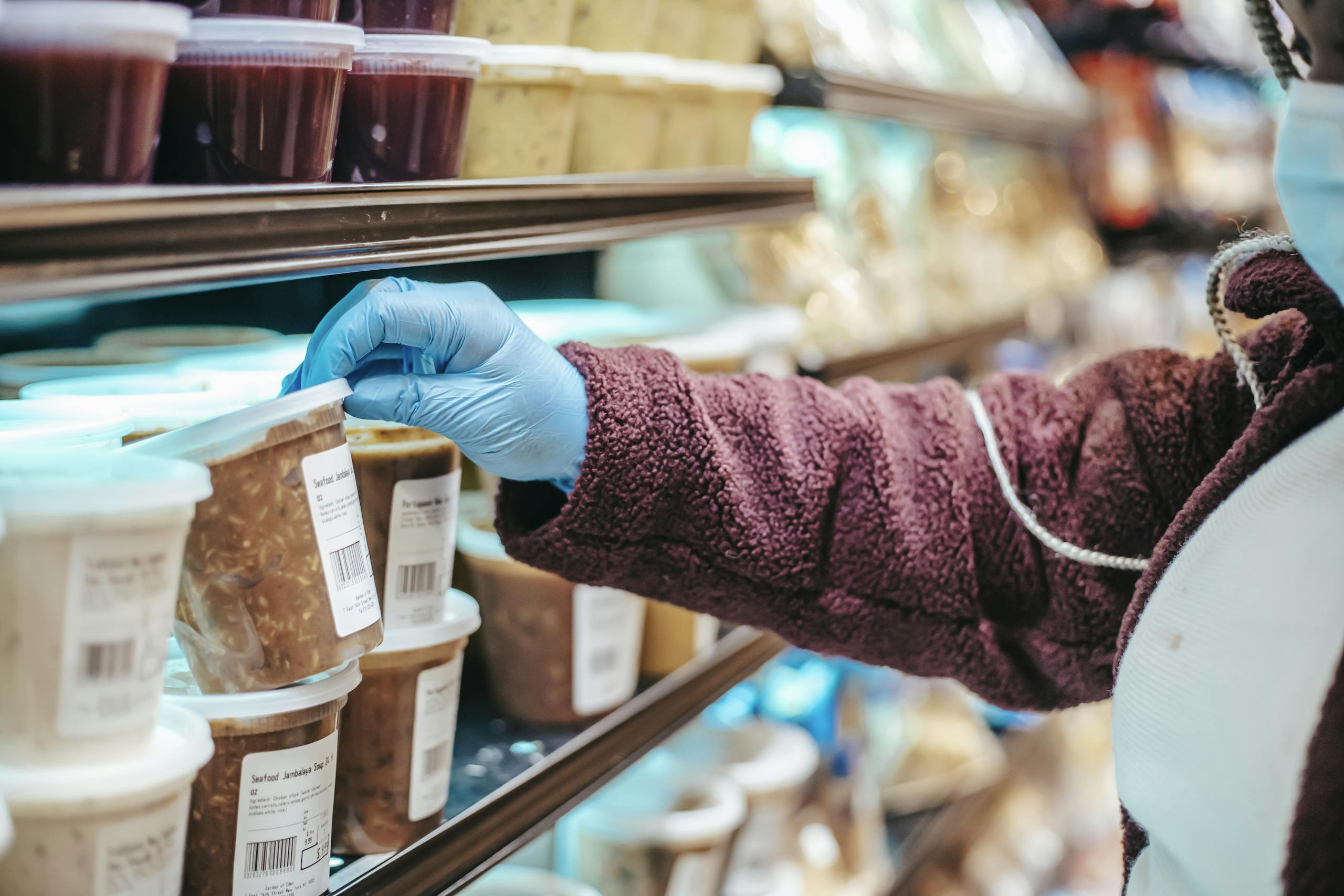 Crop anonymous female customer in protective mask reading label on frozen food in plastic container in grocery store What food recalls teach hospitality about risk management