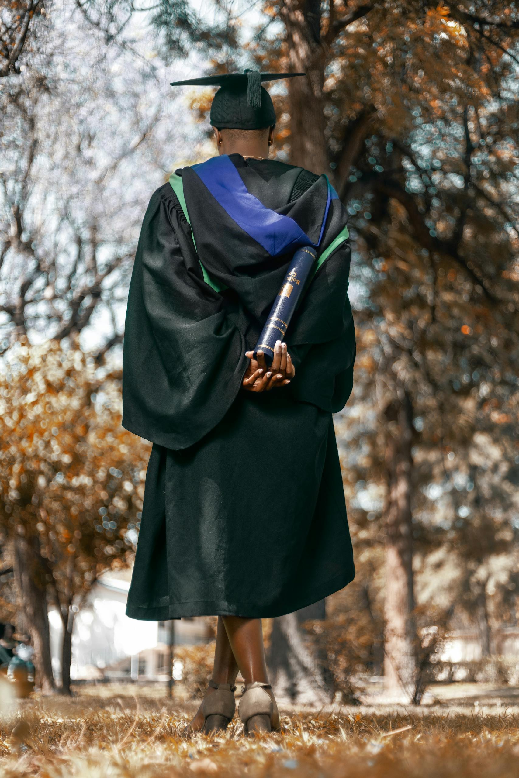 A lone graduate in a cap and gown stands in a park during fall, holding a diploma phd
