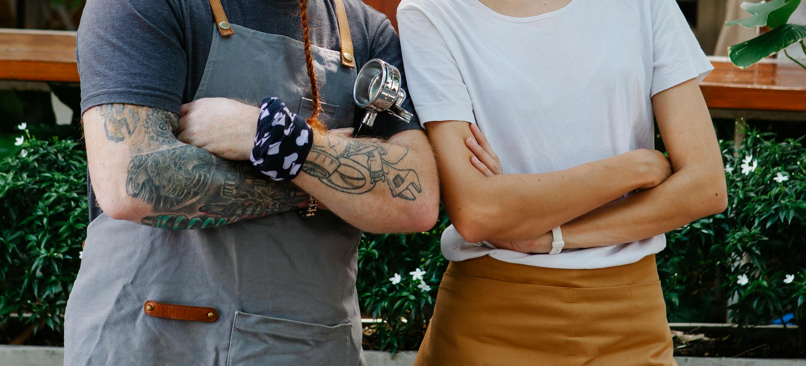 Crop anonymous couple of hospitality barista coworkers in aprons and casual clothes spending time in street with portafilter near plants in daytime
