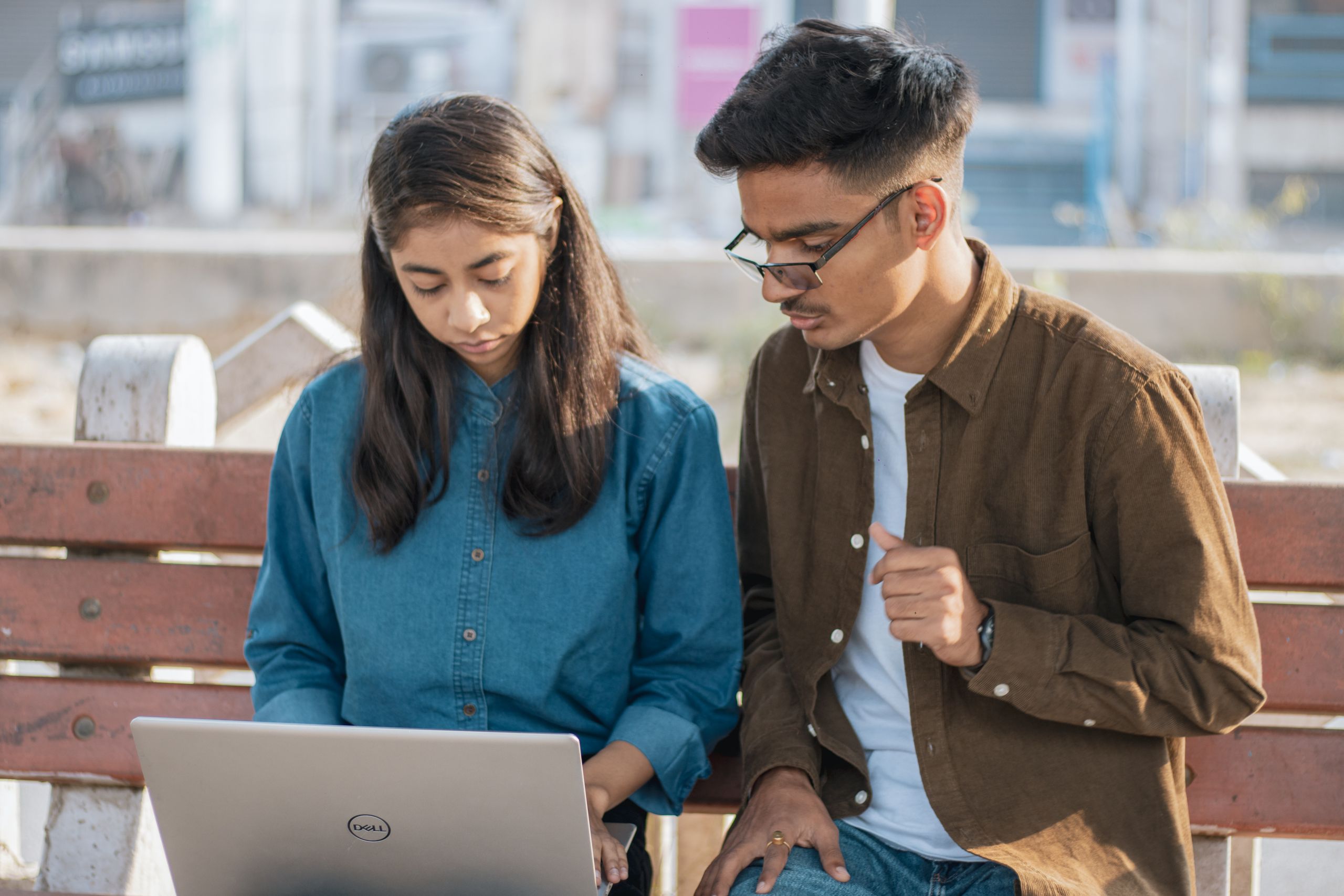 Two young adults work together on a laptop while sitting on a park bench outdoors.