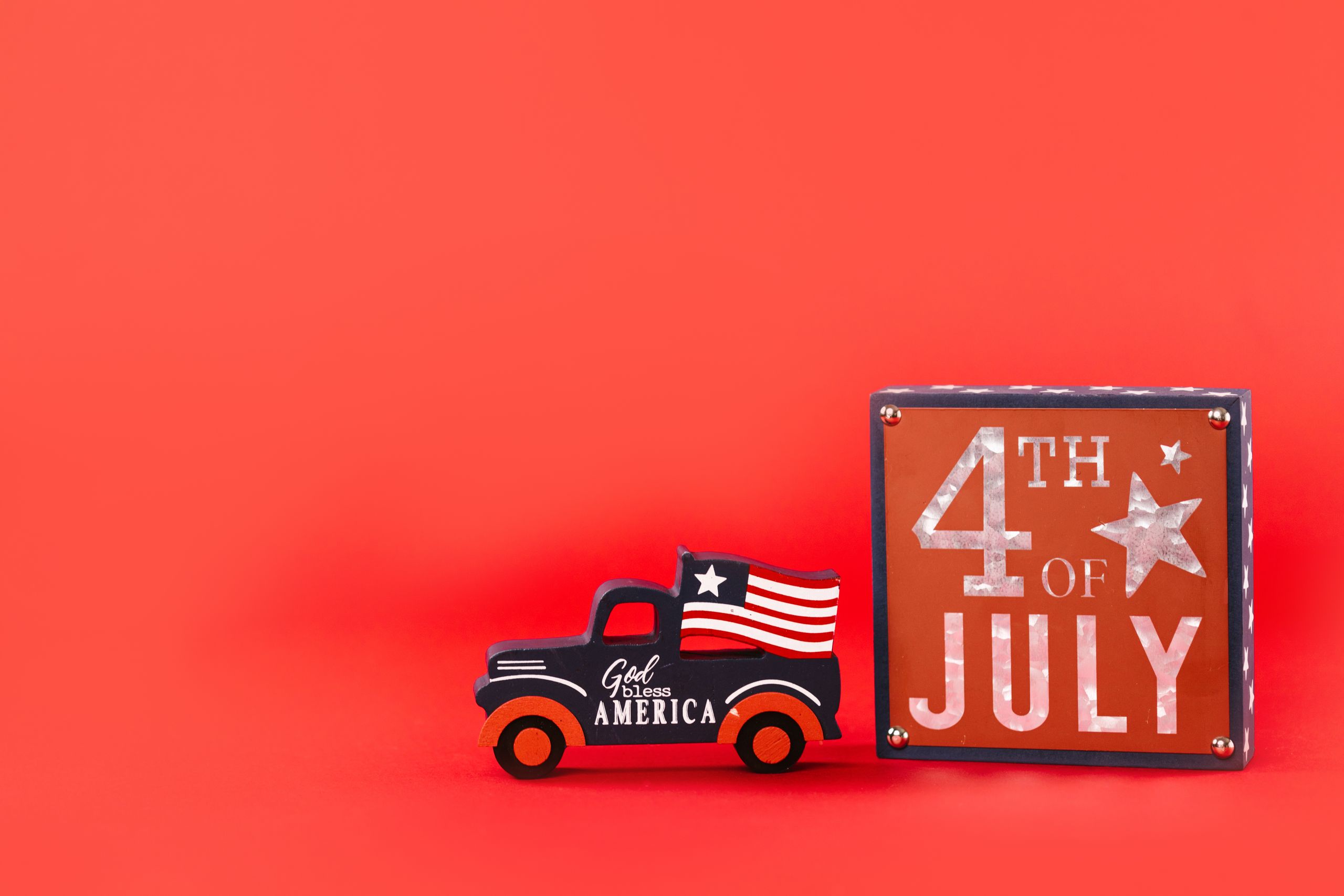 Vintage truck with American flag and 4th of July sign on red background symbolizes Independence Day celebration.