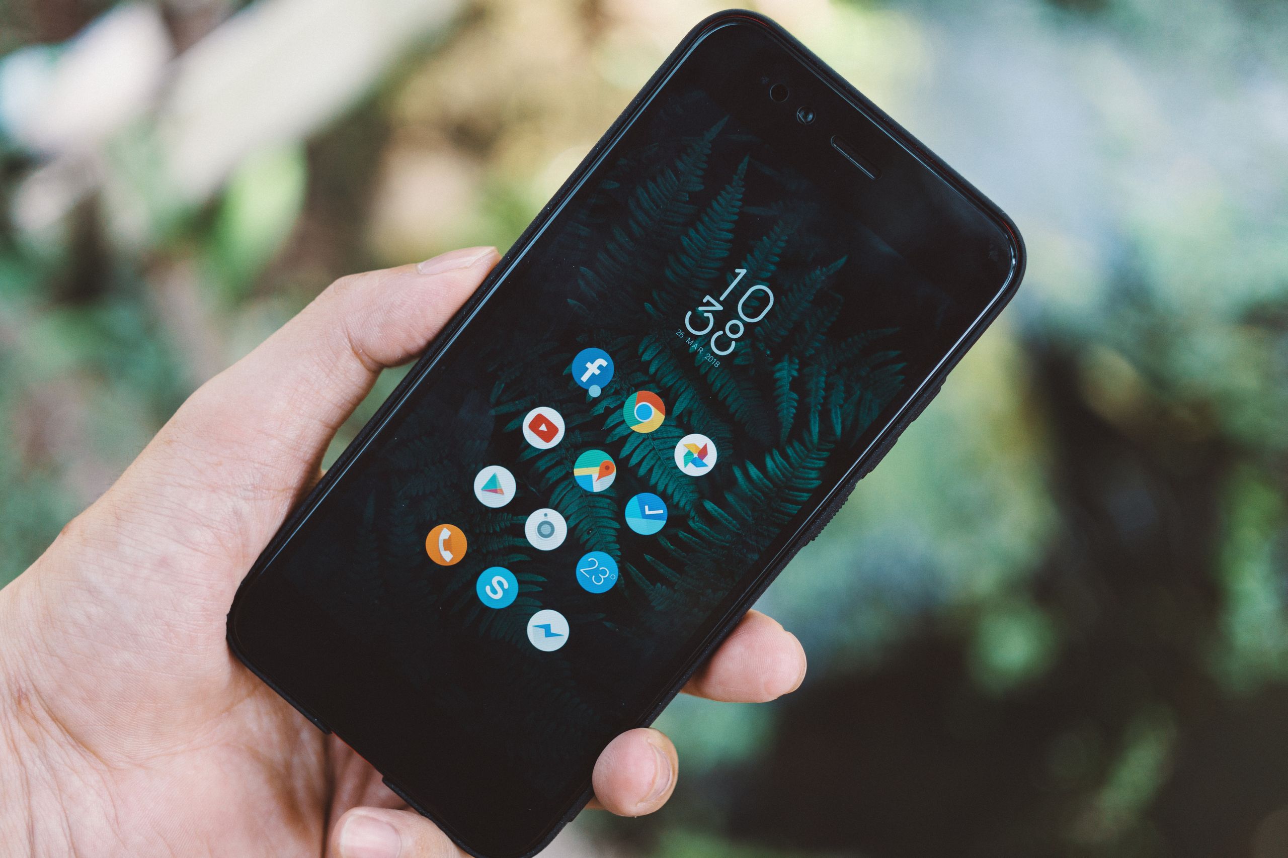 Close-up of a hand holding a smartphone with apps icons and a nature wallpaper.