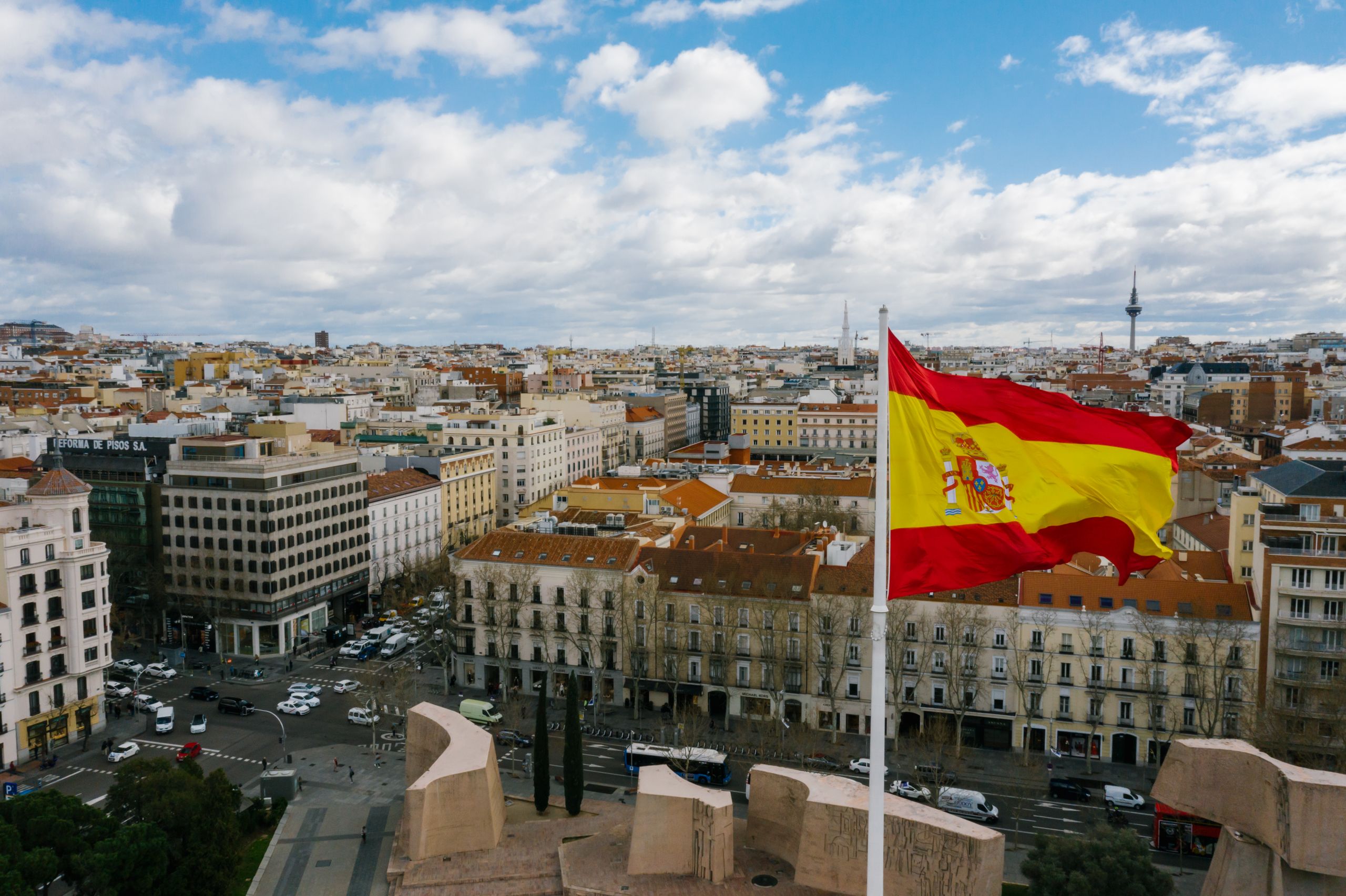 Drone view of Spanish city with aged buildings and national flag under cloudy blue sky in spain