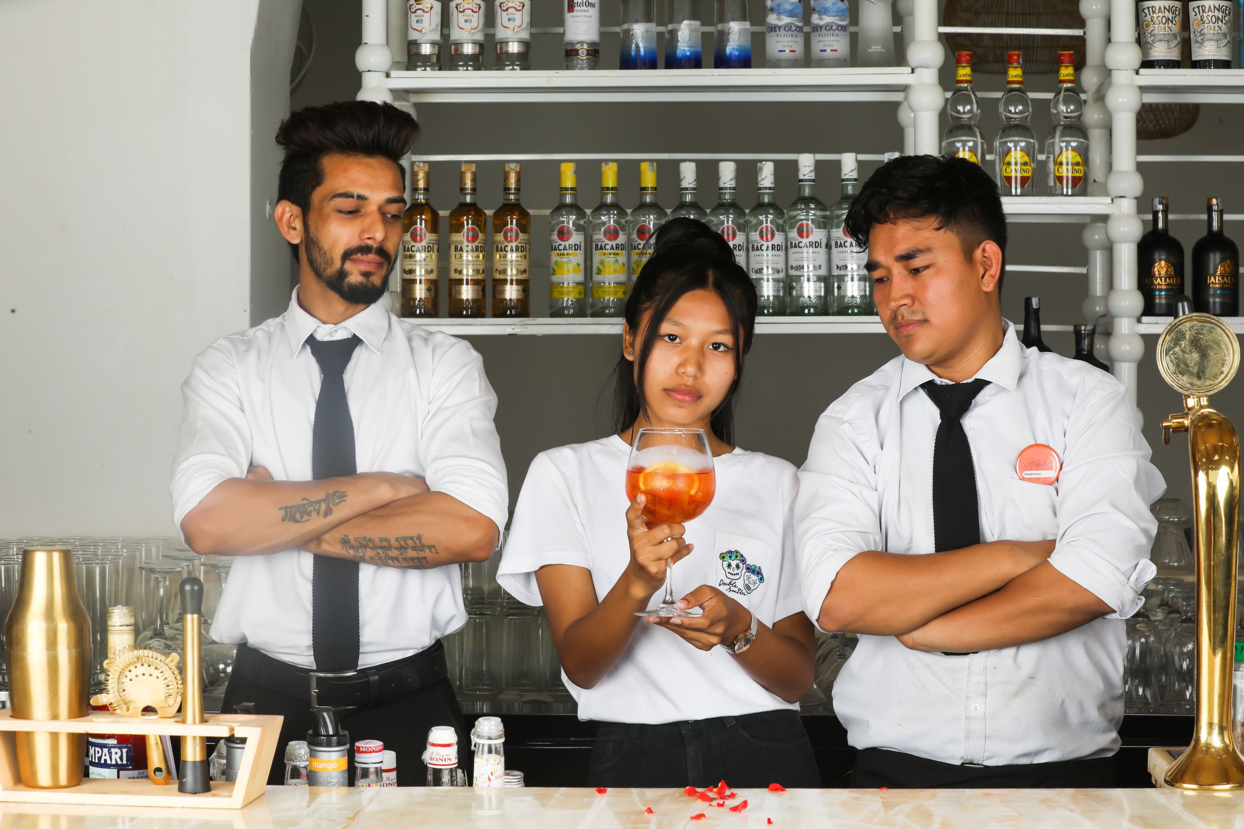 Three internship bartenders in a bar setting, discussing cocktails, surrounded by liquor bottles.