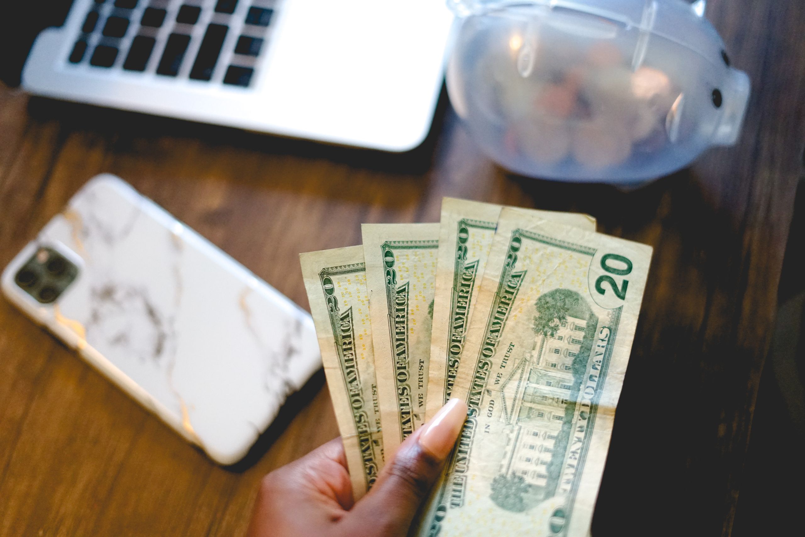 Close-up of a hand holding dollar bills beside a laptop and piggy bank, symbolizing save money and finance.